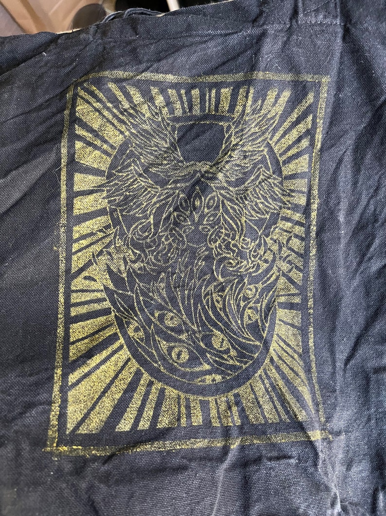 May include: Black fabric tote bag featuring a gold-coloured graphic print of a stylised winged creature with eyes. The graphic is surrounded by a sunburst design.