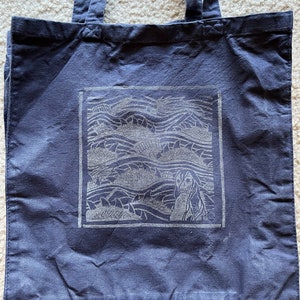May include: A dark blue canvas tote bag featuring a white graphic of a mermaid in ocean waves. The graphic is framed in a white square.