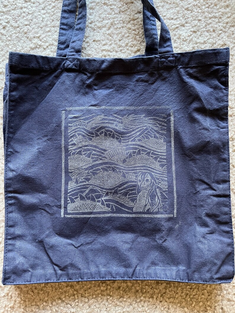 May include: A dark blue canvas tote bag featuring a white graphic of a mermaid in ocean waves. The graphic is framed in a white square.