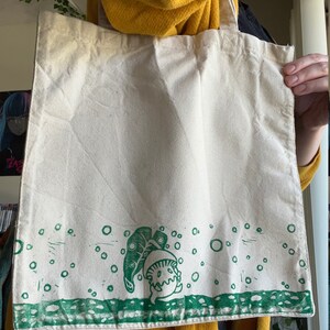 May include: A white canvas tote bag featuring a green print of a mushroom character with a leaf on top. The mushroom is surrounded by small green circles.