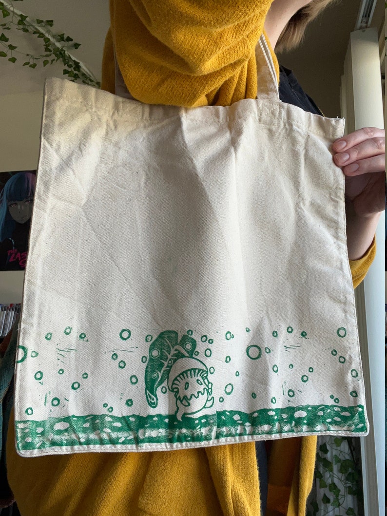 May include: A white canvas tote bag featuring a green print of a mushroom character with a leaf on top. The mushroom is surrounded by small green circles.