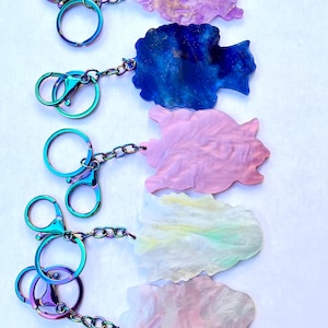 May include: Five colorful keychains with iridescent hardware. Each keychain features a unique, abstract-shaped charm in shades of purple, blue, pink, yellow, and pink. The charms are connected to the hardware with a chain.