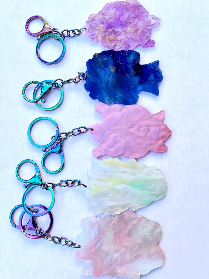 May include: Five colorful keychains with iridescent hardware. Each keychain features a unique, abstract-shaped charm in shades of purple, blue, pink, yellow, and pink. The charms are connected to the hardware with a chain.