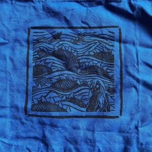 May include: Blue tote bag featuring a black and white graphic print of a wave pattern and a figure. The graphic is framed in a black rectangle.