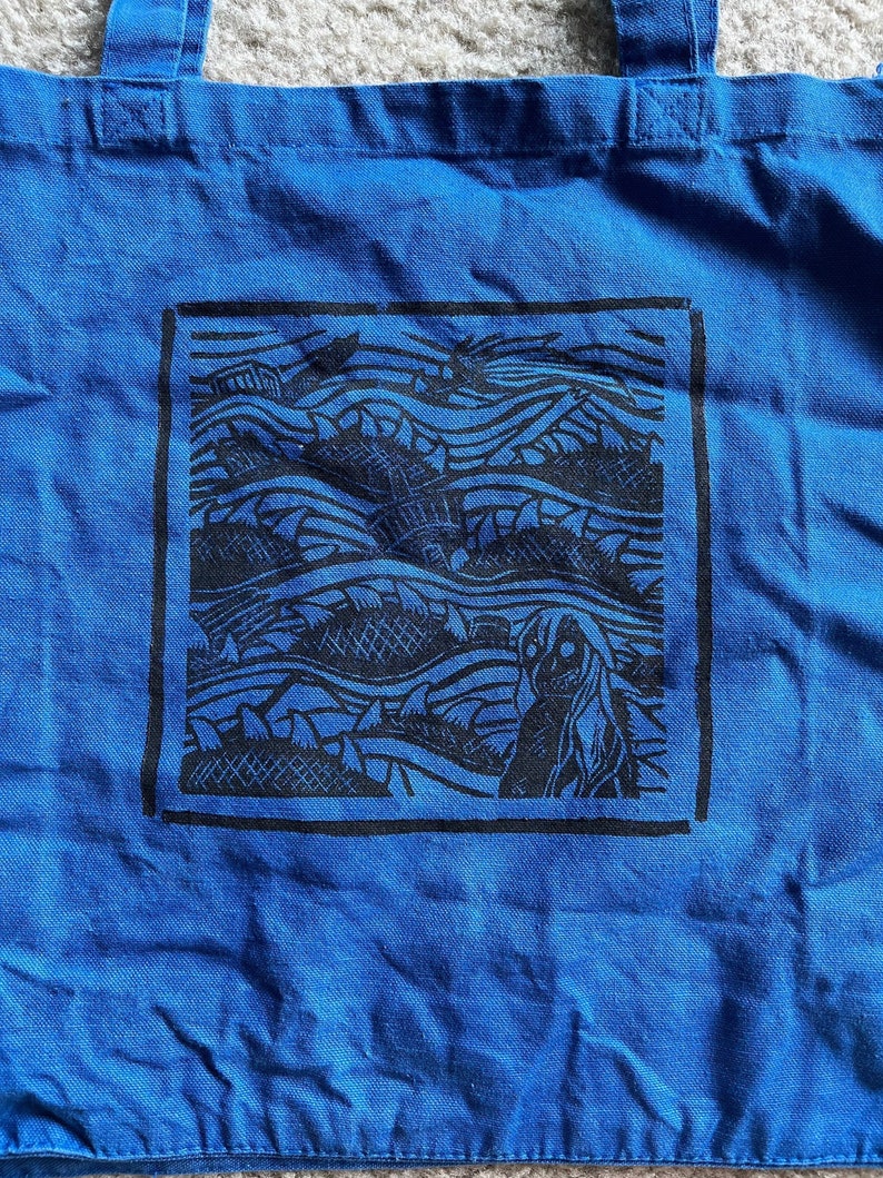 May include: Blue tote bag featuring a black and white graphic print of a wave pattern and a figure. The graphic is framed in a black rectangle.