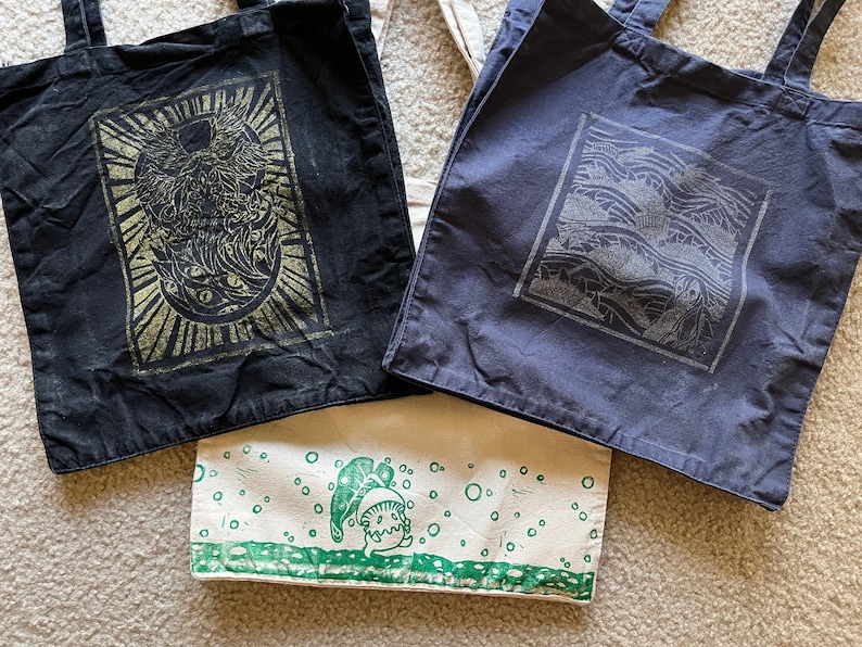 May include: Three tote bags with different designs. The first bag is black with a gold design of a creature with wings and eyes. The second bag is dark blue with a silver design of a wave pattern. The third bag is white with a green design of a creature with a leaf on its head.