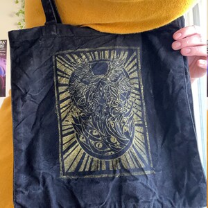 May include: Black canvas tote bag featuring a gold design of a winged creature with multiple eyes. The design is within a square frame with a sunburst pattern surrounding it.