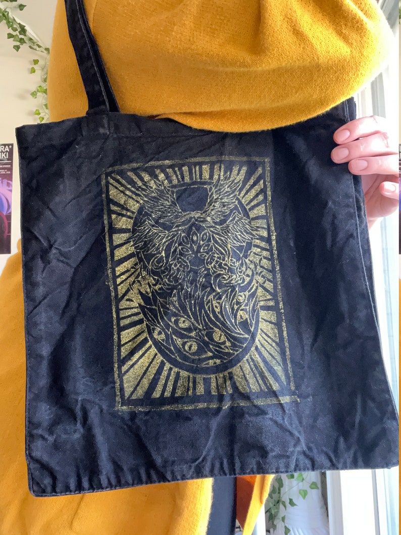 May include: Black canvas tote bag featuring a gold design of a winged creature with multiple eyes. The design is within a square frame with a sunburst pattern surrounding it.