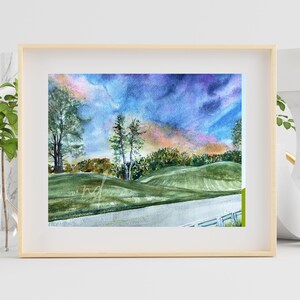 May include: Watercolor painting of a golf course landscape, framed in a light wood frame. The artwork depicts rolling green hills, trees, and a road, under a vibrant sky with shades of blue, purple, and orange. The painting has a signature in the bottom left corner.