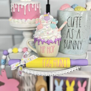 May include: A tiered tray decorated for Easter with a pink bunny cake, a white mug with a pink and purple bunny marshmallow topping, a yellow rolling pin with the text "Egg Hunt", and a blue mug with the text "Cute as a Bunny".