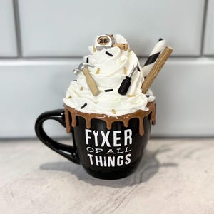 May include: Black mug with the text "Fixer of All Things" and a handle. The mug is topped with whipped cream, chocolate drizzle, and miniature tools. A hammer, screwdriver, and striped straw are visible.