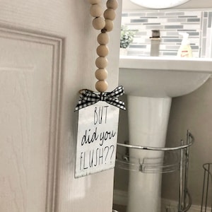 May include: A white door with a black and white checkered ribbon tied to a metal sign that reads "BUT did you FLUSH??". A wooden bead garland hangs from the door knob.