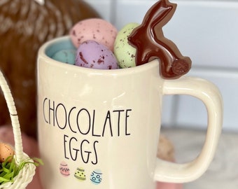Könnte beinhalten: Eine cremefarbene Tasse gefüllt mit gesprenkelten pastellfarbenen Ostereiern und einem Schokoladenhasen. Auf der Tasse steht "CHOCOLATE EGGS" mit bunten Eierillustrationen. Ein kleiner weißer Korb mit Eiern steht daneben auf einem rosa Tablett.