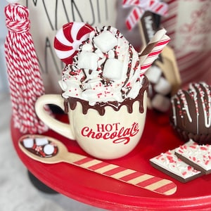 May include: A red tiered tray with a white mug filled with whipped cream, chocolate syrup, marshmallows, and red sprinkles. The mug says "Hot Chocolate". There is a candy cane, a red and white striped straw, and a wooden spoon with marshmallows on the tray.