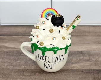 Peut inclure: Une tasse en céramique blanche avec un glaçage vert et le texte "LEPRECHAUN BAIT". La tasse est remplie de crème fouettée blanche, une saupoudrage arc-en-ciel, un pot d'or et une paille dorée.