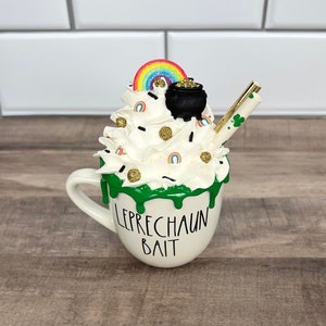 May include: White ceramic mug with green icing and the text "LEPRECHAUN BAIT". The mug is filled with white whipped cream, a rainbow sprinkle, a pot of gold, and a gold straw.