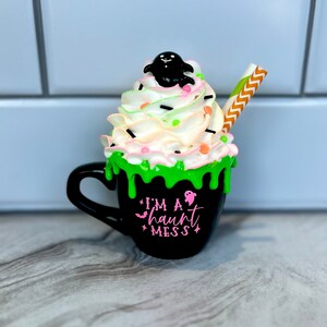 May include: Black coffee mug with green and white frosting, topped with a black ghost and sprinkles. The mug has a pink inscription that reads "I'm a haunt mess".