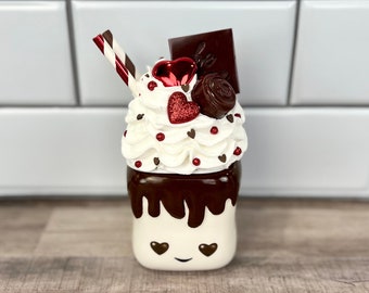 May include: A white ceramic mug shaped like a marshmallow with chocolate sauce drizzled over the top. The mug has a smiling face with two red hearts for eyes. The mug is topped with whipped cream, red candy hearts, chocolate pieces, and a red and white striped straw.