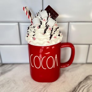 May include: Red ceramic mug with a white "COCOA" inscription. The mug is filled with whipped cream, marshmallows, chocolate pieces, and a red and white candy cane.