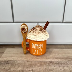 May include: Orange ceramic mug with white lettering that reads "Pumpkin Latte". The mug is filled with a white whipped cream topping and a cinnamon stick. A wooden spoon with a dollop of whipped cream is next to the mug.