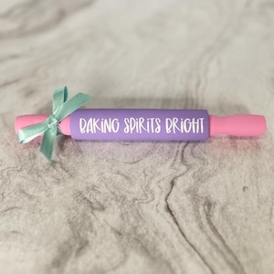 May include: Purple rolling pin with pink handles and a teal bow. The rolling pin has the text "Baking Spirits Bright" written on it.