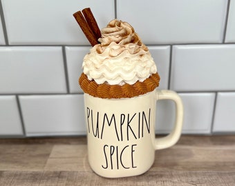 May include: A white ceramic mug with a brown rim and the words "Pumpkin Spice" written in black. The mug has a white whipped cream topping with cinnamon sprinkled on top and two cinnamon sticks sticking out of the whipped cream.