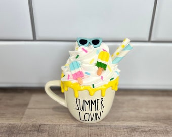 May include: White ceramic mug with yellow glaze dripping down the sides. The mug has a white whipped cream topping with colorful sprinkles, popsicles, and sunglasses. The mug says "Summer Lovin'" in black lettering.
