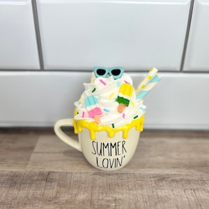 May include: White ceramic mug with yellow glaze dripping down the sides. The mug has a white whipped cream topping with colorful sprinkles, popsicles, and sunglasses. The mug says "Summer Lovin'" in black lettering.