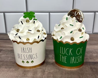 Peut inclure: Deux cupcakes blancs et verts avec une bordure dorée. Le cupcake de gauche dit "Irish Blessings." Le cupcake de droite dit "Luck of the Irish."