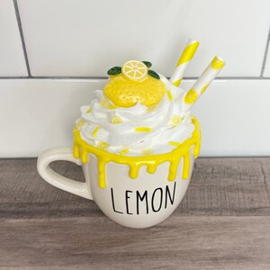 May include: White ceramic mug with a yellow lemon design and the word "LEMON" printed on the front. The mug is topped with white whipped cream and yellow lemon slices, and two yellow and white striped straws.