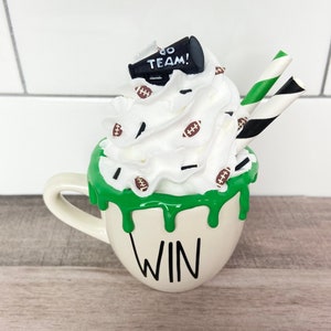 May include: White ceramic mug with green dripping glaze and the word "WIN" on the front. The mug is topped with whipped cream, chocolate sprinkles, football-shaped candies, and a miniature black megaphone with the words "GO TEAM!" on it. Two striped straws are sticking out of the whipped cream.