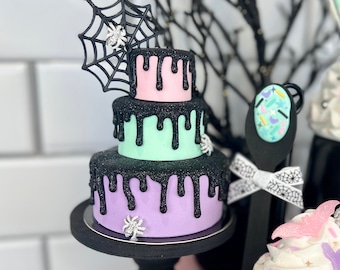 May include: A three-tiered cake decorated with black icing and purple, pink, and green layers. The cake is topped with a black spider web and a small spider. The cake is on a black pedestal and there is a small pumpkin and a cupcake in the background.