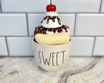 May include: A white ceramic mug with the word "SWEET" printed on the front. The mug is filled with a chocolate and vanilla ice cream sundae topped with a cherry, whipped cream, chocolate sauce, and sprinkles.
