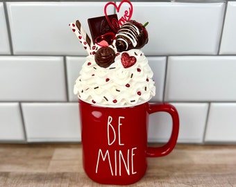 May include: A red mug with the words "BE MINE" in white, filled with whipped cream and topped with chocolate-covered strawberries, chocolate pieces, and a heart-shaped decoration. The mug is on a wooden surface.