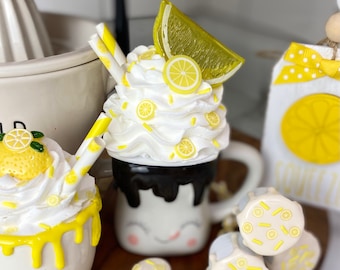 May include: A white mug with a black glaze and a happy face design, topped with whipped cream and lemon slices. The mug is surrounded by yellow and white lemon-themed marshmallows and a smaller mug with a yellow glaze and lemon slices.