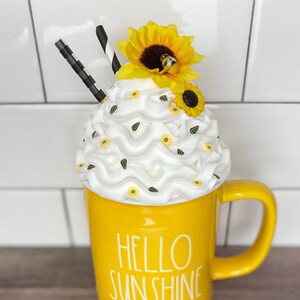May include: Yellow ceramic mug with the text "Hello Sunshine" in white lettering. The mug is topped with white whipped cream and a yellow sunflower with a small bee.