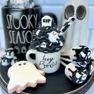 May include: A black and white Halloween themed mug with the text "hey boo!" and a ghost-shaped marshmallow on top. The mug is surrounded by other Halloween decorations, including a ghost cookie, a ghost-shaped mug with the text "Spooky Season", and a ghost-shaped marshmallow on a stick.