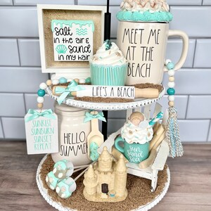 May include: A two-tiered white tiered tray decorated with a beach theme. The top tier features a white mug with the text "Meet me at the beach", a cupcake with turquoise frosting, a wooden sign that says "Life's a beach", and a small jar with a turquoise lid. The bottom tier features a white mug with the text "Sun sand sea", a small sandcastle, a jar with the text "Hello Summer", and a sign with the text "Sunrise, sunburn, sunset, repeat".