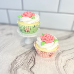 May include: Two cupcakes with white frosting and pink rose decorations. The cupcakes are in floral patterned paper liners. The cupcakes are on a white cake stand.