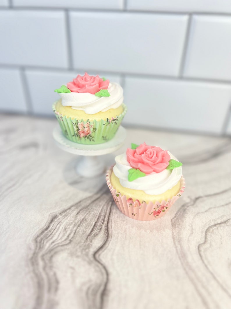 May include: Two cupcakes with white frosting and pink rose decorations. The cupcakes are in floral patterned paper liners. The cupcakes are on a white cake stand.