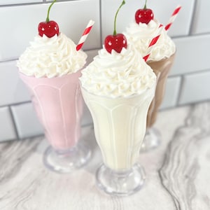 May include: Three milkshakes with whipped cream and a cherry on top. The milkshakes are in clear glass mugs with a red and white striped straw in each. The milkshakes are pink, white, and brown.