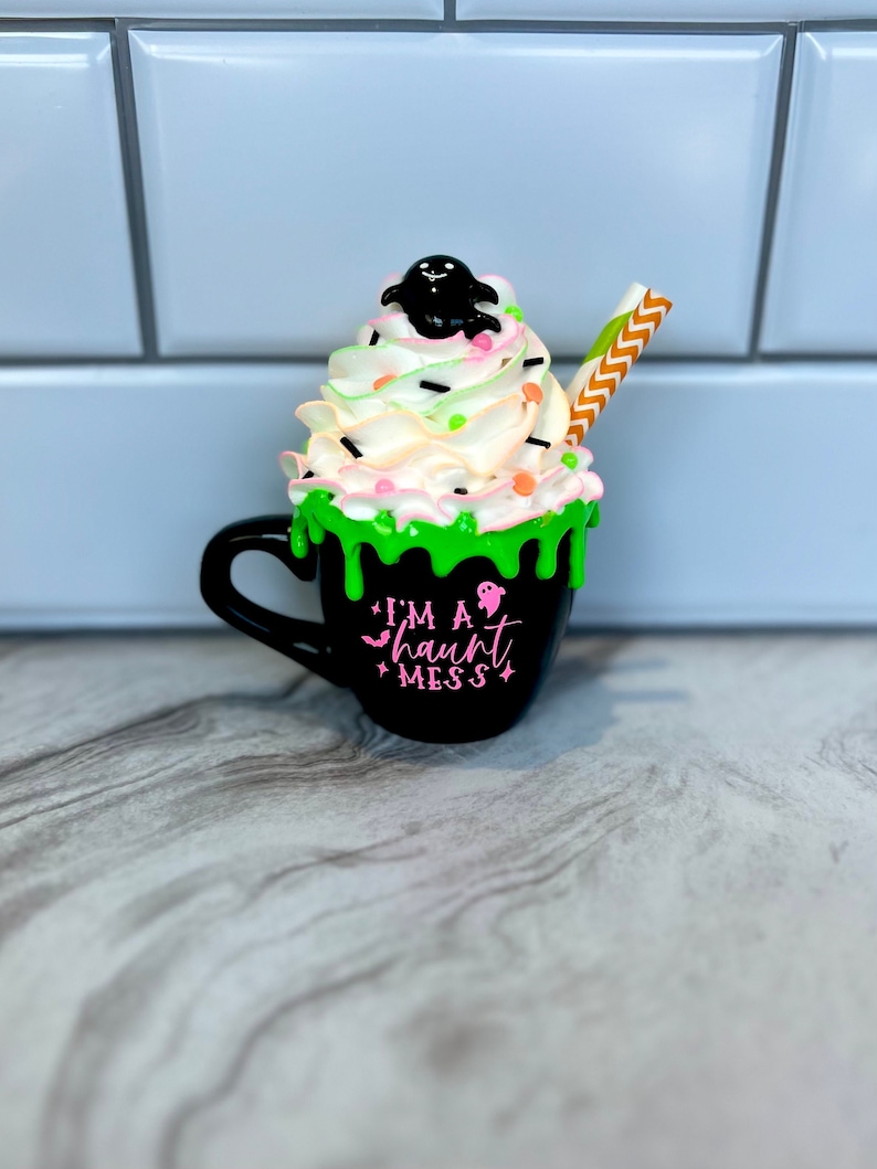 May include: Black coffee mug with green and white frosting, topped with a black ghost and sprinkles. The mug has a pink inscription that reads "I'm a haunt mess".