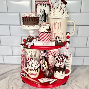 May include: A two-tiered red tiered tray decorated with a hot chocolate theme. The top tier features a white mug with the words "HOT COCOA" in red, a chocolate cake, a cupcake with red and white stripes, and a sign that says "Hot Cocoa Bar". The bottom tier features a white mug with the words "HOT CHOCOLATE" in red, a white mug with a smiley face, a white mug with the words "Oh What Fun", a white mug with the words "Marshmallow", and a sign that says "Cocoa Ags: Marshmallows, Whipped Cream, Canes".