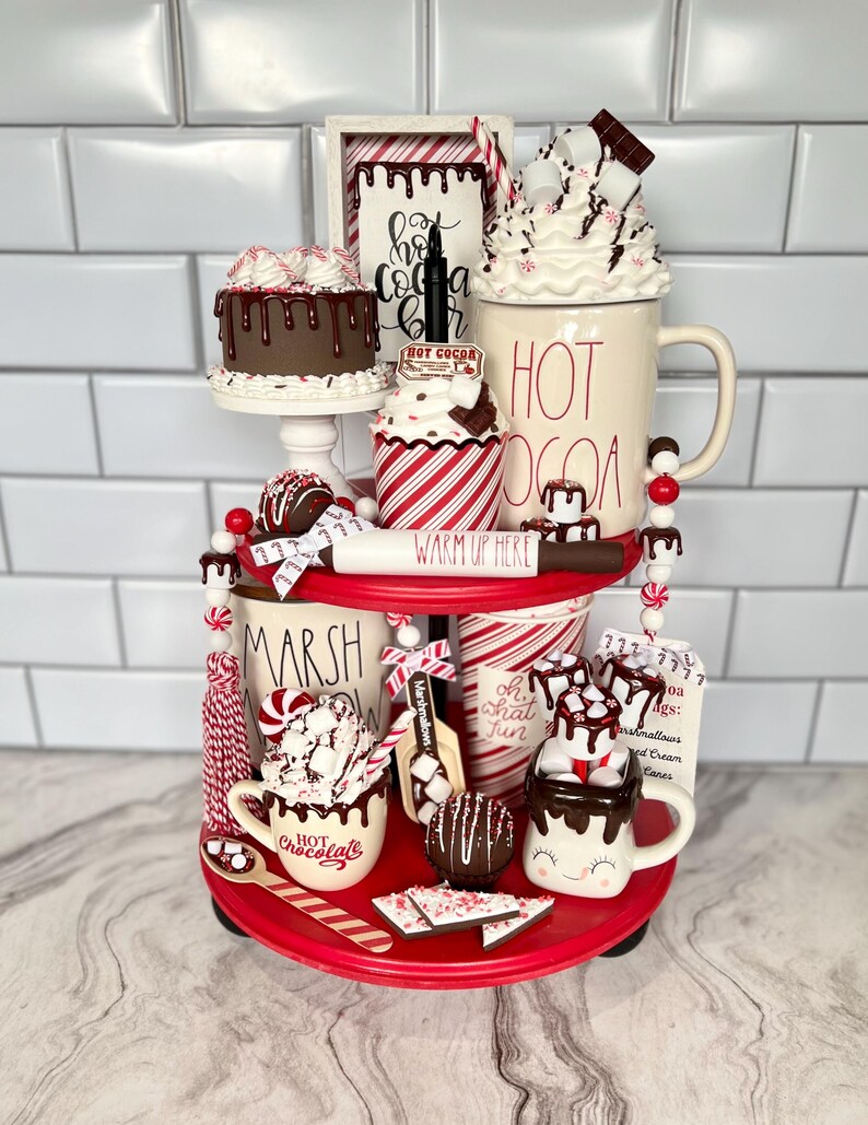 May include: A two-tiered red tiered tray decorated with a hot chocolate theme. The top tier features a white mug with the words "HOT COCOA" in red, a chocolate cake, a cupcake with red and white stripes, and a sign that says "Hot Cocoa Bar". The bottom tier features a white mug with the words "HOT CHOCOLATE" in red, a white mug with a smiley face, a white mug with the words "Oh What Fun", a white mug with the words "Marshmallow", and a sign that says "Cocoa Ags: Marshmallows, Whipped Cream, Canes".