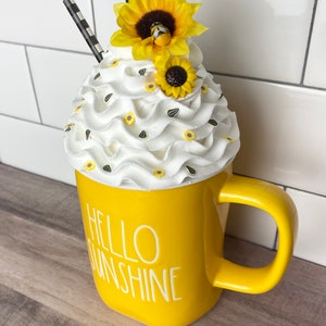 May include: Yellow ceramic mug with the text "Hello Sunshine" and a white whipped cream topping decorated with small yellow flowers and a bee.