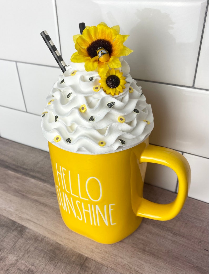 May include: Yellow ceramic mug with the text "Hello Sunshine" and a white whipped cream topping decorated with small yellow flowers and a bee.