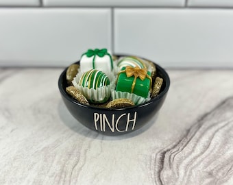 Peut inclure: Un bol noir avec le mot "PINCH" sur le devant, rempli de bonbons au chocolat vert et blanc décorés de nœuds dorés. Le bol est posé sur une surface blanche.