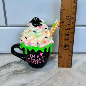 May include: A black ceramic mug with green icing and white whipped cream topped with a black ghost and colorful sprinkles. The mug has the text "I'm a haunt mess" in pink lettering.