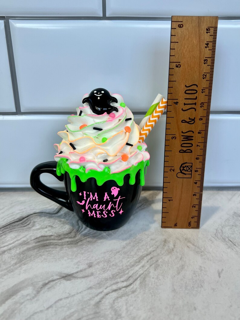 May include: A black ceramic mug with green icing and white whipped cream topped with a black ghost and colorful sprinkles. The mug has the text "I'm a haunt mess" in pink lettering.