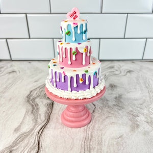 May include: A three-tiered cake decorated with colorful candy hearts and white frosting. The top tier is light blue, the middle tier is pink, and the bottom tier is purple. The cake is on a pink cake stand. The top of the cake has a candy heart that says "Hug Me".